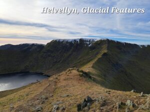 A view from a grassy/rocky area along a ridge leading to a longer wider ridge which is Helvellyn mountain. On the right the ridge drops away in a grassy slope and on the left the slope drops away to a Glacial pool. There are traces of snow on the main ridge. The sky is mainly thin cloud with some blue sky on the right. On the left the sky has a pink tinge from a weak sun.