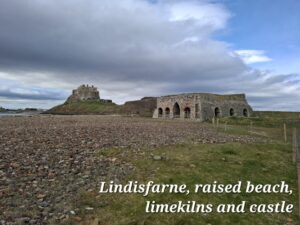 Looking across a pebbly beach on the left and a grassy area on the right. Beyond this you can see brick built lime kilns with seven arched entrances. Further back is Lindisfarne Castle sat on top of a small conical hill. There is blue sky to the right and a large grey cloud over the Castle.