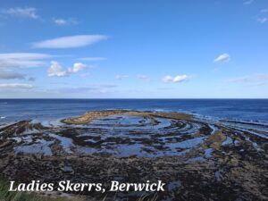 Looking at a beach with a circle of rocks round a rock pool of water. Beyond this is a deep blue sea and a blue sky with a few small white clouds.