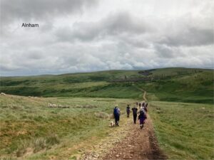 A group of around ten people and a dog wlaking away down a rock path with grass on both sides and a hill in front of them. there are grey clouds overhead.
