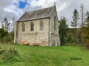 A small rectangular church with just three windows on the side and one at the end. It is set in a grassy area with a few trees behind it. The sky is cloudy with one small patch of blue above and to the left of the church.