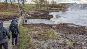 A group of about six walkers approaching and crossing a footbridge with the sea breaking over rocks, with lots of spray on the right hand side. There is some blue sky and white clouds just visible through bare trees at the top of the scene