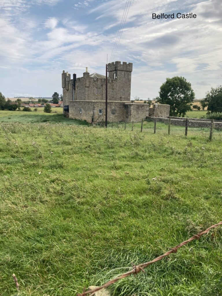 Looking across some rough grass to a small square castle with a small tower with battlements on top. The sly is blue and grey with some white patches of cloud.