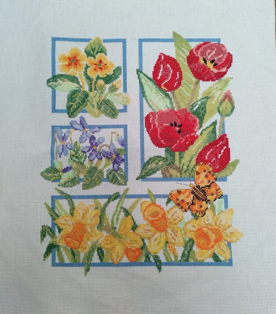 A cross stitch picture made up of blue bordered sections containing primroses, violets, Tulips and daffofils with an orange butterfly flying over.