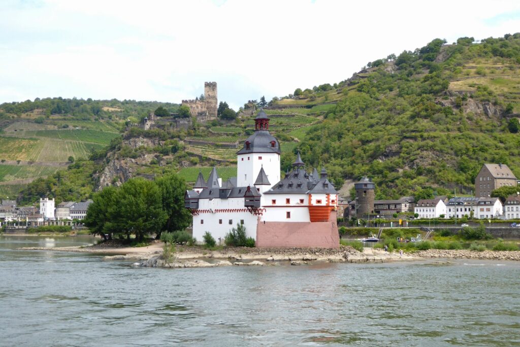 Looking over the water from a boat. There is a castle on the hill on the left and you can see part of a village along the Riverside. A white castle on the edge of the river is in the foreground. There is a green hillside behind and a slightly cloudy sky.